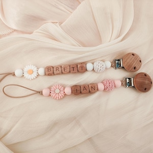 May include: Two baby pacifier clips with wooden beads spelling out names. One clip features a daisy and the other a pink flower. The clips have wooden and crochet beads, and wooden round clips.