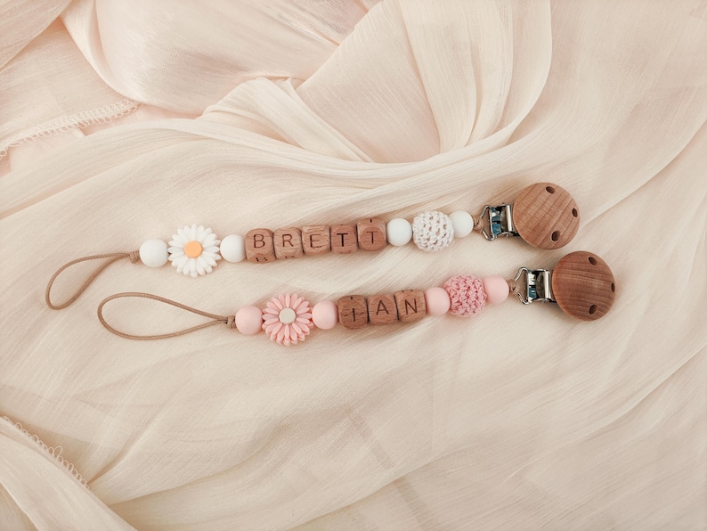 May include: Two baby pacifier clips with wooden beads spelling out names. One clip features a daisy and the other a pink flower. The clips have wooden and crochet beads, and wooden round clips.