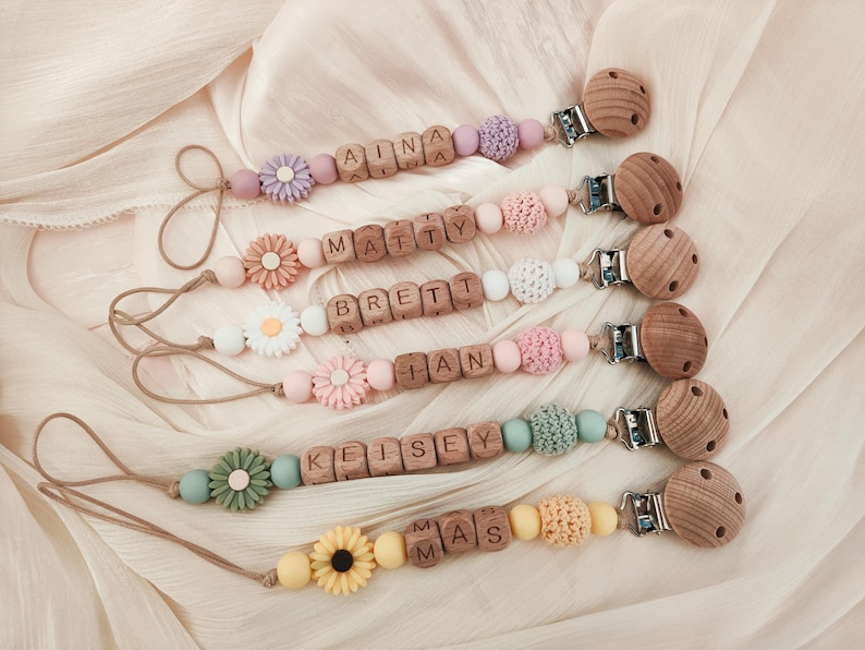 May include: Six personalized baby pacifier clips with wooden beads spelling out names. Each clip features a wooden clip, a wooden bead, and decorative elements like flowers and crochet beads in pastel colors.
