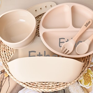 May include: A beige baby feeding set, including a divided plate, bowl, bib, and utensils. The plate and utensils are personalized with the name "Ethan". The set is made of silicone and wood.