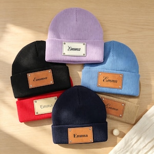 May include: A collection of six knit beanies in lavender, black, red, blue, tan, and navy blue. Each beanie features a rectangular patch attached with four metal rivets. The patches are engraved with the name Emma in a cursive script font.