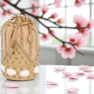 Könnte beinhalten: Eine goldfarbene Beuteltasche mit Perlenakzenten und Muscheldetails. Die Tasche hat ein dekoratives Muster und einen Kordelzugverschluss. Rosa Blütenblätter sind auf der Oberfläche verstreut, mit einem verschwommenen floralen Hintergrund.
