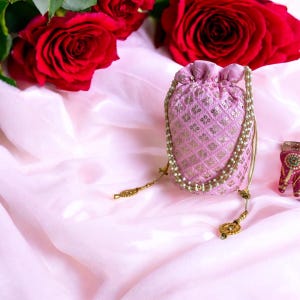 Könnte beinhalten: Ein rosa Kordelzugbeutel mit goldenen Verzierungen und einem Perlenriemen liegt auf einem rosa Stoff. Rote Rosen sind im Hintergrund. Die Tasche hat ein Blumenmuster und einen goldenen Kordelzugverschluss.