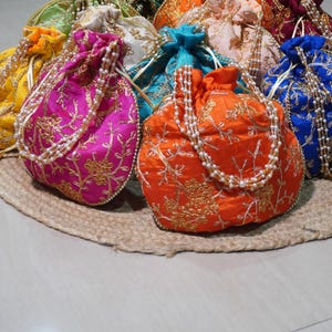 Puede incluir: Una colección de coloridas bolsas con cordón bordadas en varios tonos, incluyendo rosa, naranja y azul. Cada bolsa está adornada con hilo dorado y un asa de cuentas, descansando sobre una estera tejida. Las bolsas son probablemente para llevar objetos pequeños.