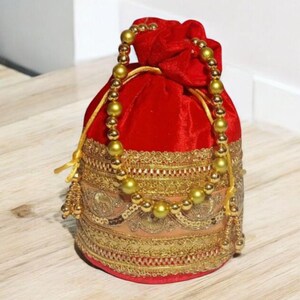 Könnte beinhalten: Ein roter Samtbeutel mit Kordelzug, goldfarbenem Perlenhenkel und dekorativen goldfarbenen Zierleisten. Die Tasche hat aufwendige Stickereien und goldfarbene Quasten. Die Tasche steht auf einer hellen Holzoberfläche.