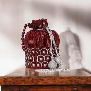 Op de afbeelding: Een bordeauxrode, met kralen bezette trekkoordtas met een kralen handvat en zilveren kwastjes. De tas heeft een decoratief patroon van witte parels en zilveren accenten rond de basis. De tas staat op een houten oppervlak.
