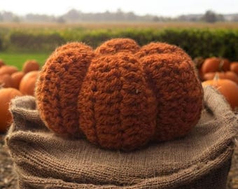 Pode incluir: Uma abóbora laranja de crochê feita à mão com uma superfície texturizada, repousando sobre um saco de serapilheira. A abóbora é de uma cor outonal quente, e outras abóboras são visíveis no fundo, sugerindo um tema de colheita de outono.