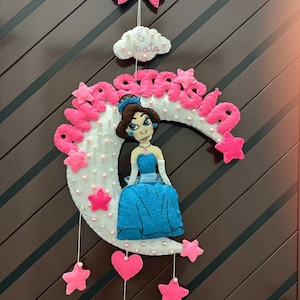 Può includere: Un'applique da parete a tema principessa in feltro rosa, bianco e blu. Il design include una luna crescente, stelle, cuori e una figura di principessa. Il nome "Anastasia" è scritto in lettere rosa. Un fiocco rosa e una nuvola con il nome "Naia" sono in alto.