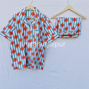 May include: A two-piece pajama set with a white background, featuring vertical teal stripes and orange sunburst patterns. The set includes a button-down shirt with a chest pocket and matching elastic-waist pants.