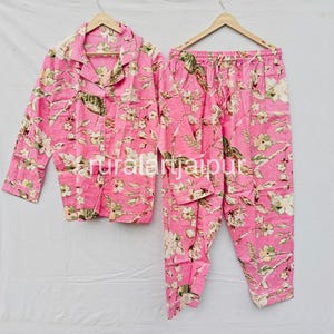 May include: Pink floral pajama set with a button-down top and matching pants. The set features a vibrant floral pattern in shades of white, green, and yellow. The long-sleeved top has a collared neckline and a breast pocket. The pants have a drawstring waist.