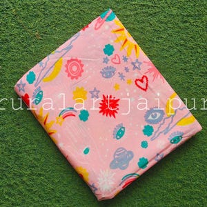 May include: A folded piece of light pink fabric featuring a repeating pattern of yellow suns, red hearts, blue eyes, rainbows, planets, and white stars. The text "ruralartjaipur" is printed in white across the centre of the fabric.