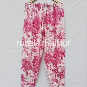 May include: Pink and white pajama pants with an elastic waistband, hanging on a wooden hanger. The pants feature a repeating pattern of tropical plants, birds, and tigers. The text "ruralartjaipur" is printed on the pants.