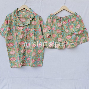 May include: A two-piece pyjama set with a floral pattern. The set includes a short-sleeved button-down shirt and matching shorts. The fabric is a teal colour with pink and yellow floral designs. The shirt and shorts are hanging on wooden hangers.