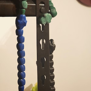 May include: A handmade beaded pendant with a blue skull and cube design, transitioning to green cube beads. The pendant is hanging on a dark metal structure. The beads are made of a solid material.