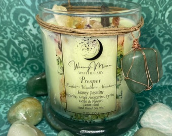 May include: A glass candle jar with a brown label that reads "Waxing Moon Apothecary Prosper Health Wealth Abundance Honey Jasmine Citrine, Green Aventurine, Pyrite Herbs & Flowers Custom Blend Hand Poured Soy Wax 9 oz/255 g". The candle jar is surrounded by green stones and has a green stone charm hanging from the top.
