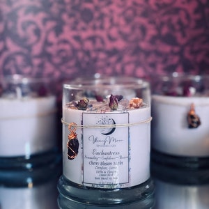 May include: A glass candle jar with a white label that reads "Woxing Moon Apothecary Enchantress Sensuality Confidence Passion Cherry Blossom Merlot Carnelian, Garnet, Herbs & Flowers Custom Blend Hand Poured Soy Wax". The jar is decorated with dried flowers and a small crystal pendant.