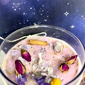 May include: A pink candle with dried flowers, herbs, and crystals. The candle is in a clear glass jar with a white label.