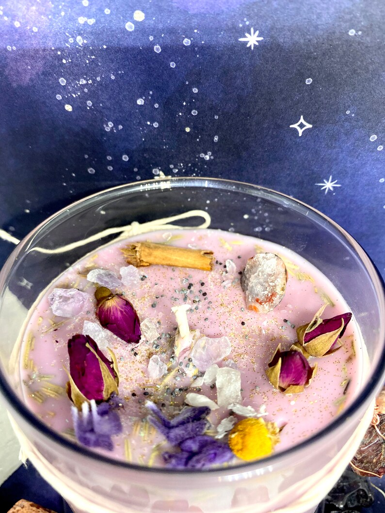 May include: A pink candle with dried flowers, herbs, and crystals. The candle is in a clear glass jar with a white label.