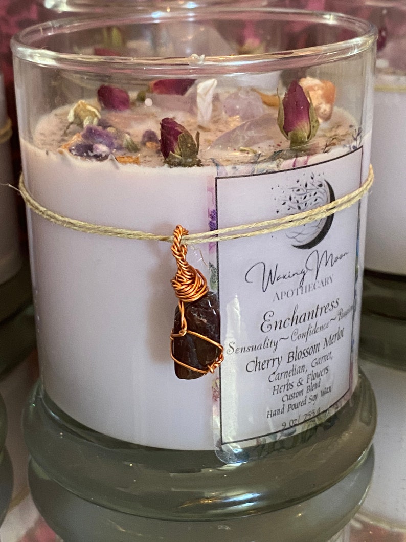 May include: A glass candle jar containing a white soy wax candle. The candle is decorated with dried flowers, crystals, and a copper wire wrapped garnet stone. The label reads "Waxing Moon Apothecary Enchantress Sensuality~Confidence Cherry Blossom Merlot Carnelian, Garnet, Herbs & Flowers Custom Blend Hand Poured Soy Wax 9 oz/255.4"