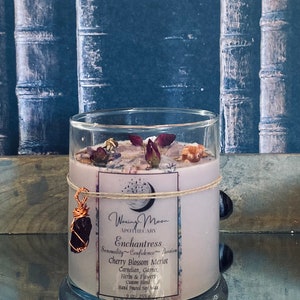 May include: A glass candle jar with a pink label that reads "Waxing Moon Apothecary Enchantress Sensuality Confidence Passion Cherry Blossom Merlot Carnelian, Garnet, Herbs & Flowers Custom Blend Hand Poured Soy Wax 9.0 oz/255g". The candle jar is decorated with dried flowers and a copper wire wrapped gemstone.