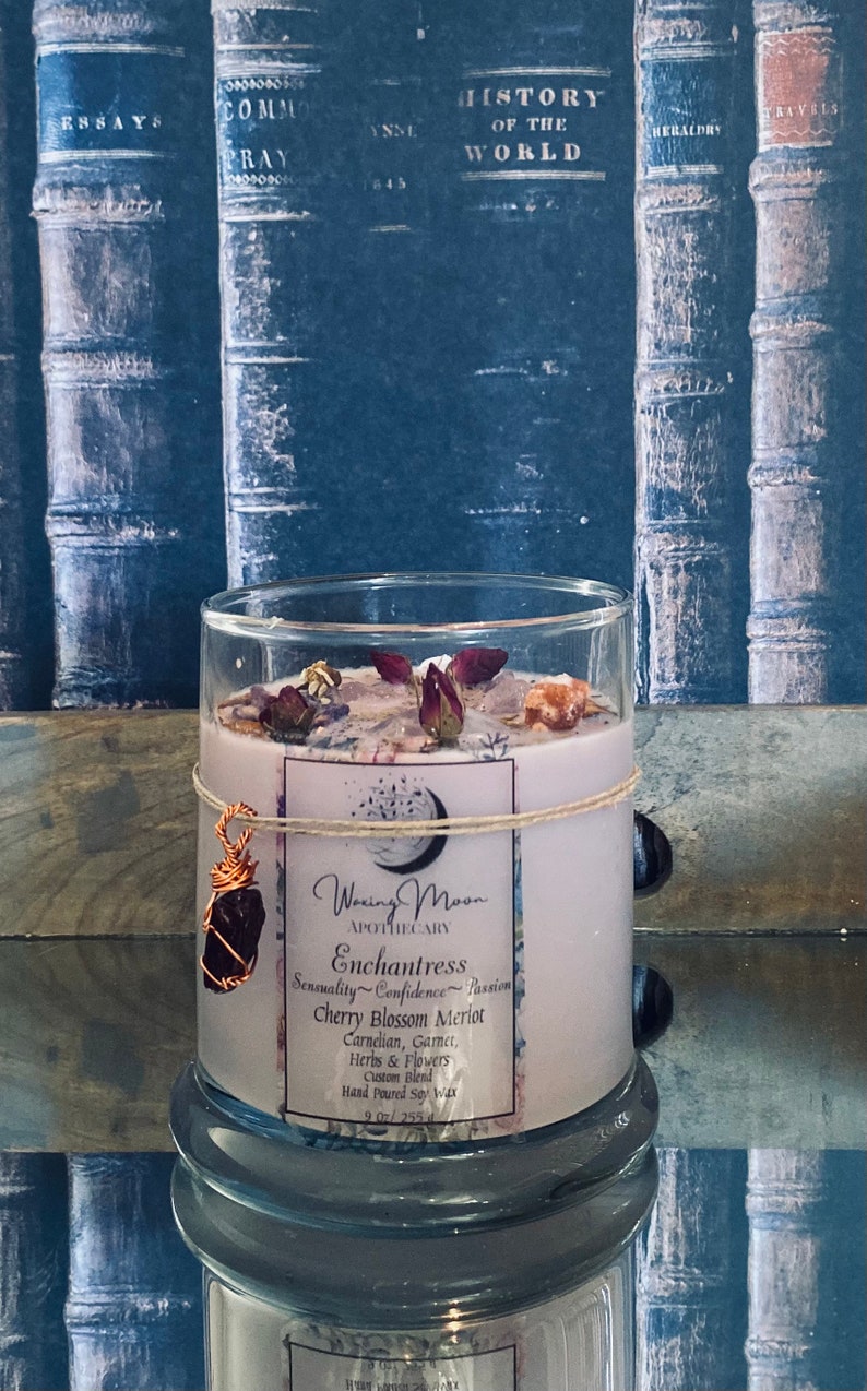May include: A glass candle jar with a pink label that reads "Waxing Moon Apothecary Enchantress Sensuality Confidence Passion Cherry Blossom Merlot Carnelian, Garnet, Herbs & Flowers Custom Blend Hand Poured Soy Wax 9.0 oz/255g". The candle jar is decorated with dried flowers and a copper wire wrapped gemstone.