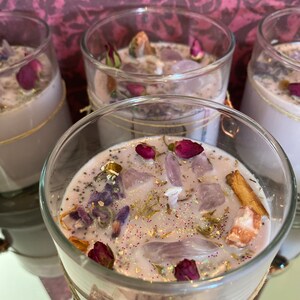 May include: Four glass candle jars containing a light purple wax base. The jars are filled with assorted dried flowers, crystals, and glitter. The candles are labelled "Wisteria Moon".