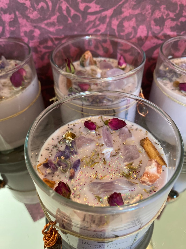 May include: Four glass candle jars containing a light purple wax base. The jars are filled with assorted dried flowers, crystals, and glitter. The candles are labelled "Wisteria Moon".