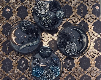 May include: Four black and silver round candle holders with a moon, face, and flower designs. The candle holders are on a black and gold patterned background.