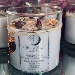May include: A white soy candle in a clear glass jar with a teal blue base. The candle is decorated with dried flowers and crystals. The label reads "Woxing Moon Apothecary Enchantress Sensuality Confidence Passion Cherry Blossom Merlot Carnelian, Garnet, Herbs & Flowers Custom Blend Hand Poured Soy Wax 9.02/255g"