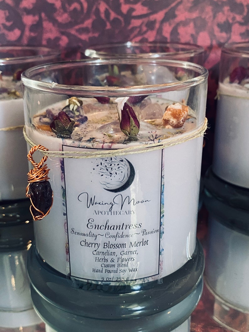 May include: A white soy candle in a clear glass jar with a teal blue base. The candle is decorated with dried flowers and crystals. The label reads "Woxing Moon Apothecary Enchantress Sensuality Confidence Passion Cherry Blossom Merlot Carnelian, Garnet, Herbs & Flowers Custom Blend Hand Poured Soy Wax 9.02/255g"