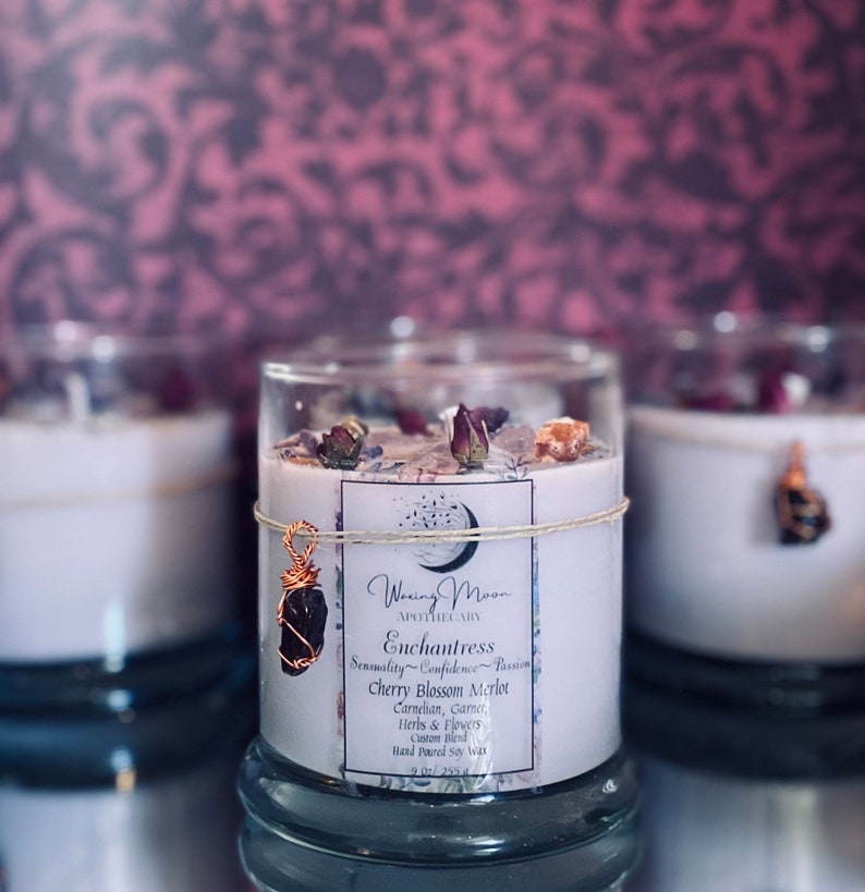 May include: A glass candle jar with a white label that reads "Woxing Moon Apothecary Enchantress Sensuality Confidence Passion Cherry Blossom Merlot Carnelian, Garnet, Herbs & Flowers Custom Blend Hand Poured Soy Wax". The candle jar is decorated with dried flowers and a small crystal pendant.