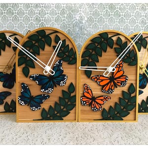 Peut inclure: Horloges murales en bois avec des motifs de papillons et de feuilles. Les horloges présentent un fond en bois clair, une bordure dorée et des aiguilles blanches. Les papillons sont peints dans des tons de bleu et d'orange. Des feuilles vertes sont également incluses.
