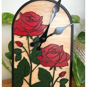 Peut inclure: Une horloge décorative en bois avec un motif floral de roses rouges et de feuilles vertes. L'horloge a un cadre noir et des aiguilles noires. Le fond est un grain de bois clair.