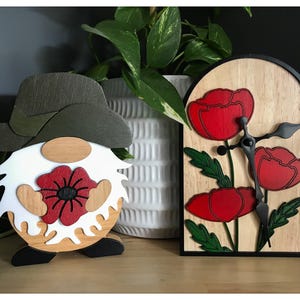 Peut inclure: Figurine de gnome en bois avec un chapeau vert, une barbe blanche et un coquelicot rouge. À côté, une horloge en bois avec des coquelicots rouges, des aiguilles noires et un cadre noir. Articles décoratifs.