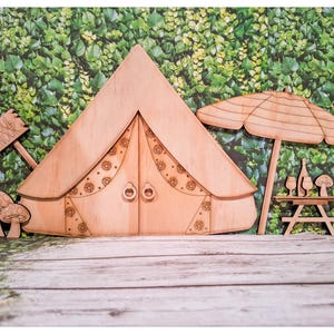 May include: Wooden fairy garden set featuring a tent, picnic table with wine and glasses, mushrooms, and an umbrella. The tent has decorative floral patterns and the text "SOUL NOOK" on a sign. The set is made of light brown wood.