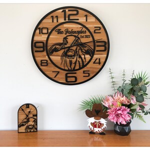 Peut inclure: Une grande horloge murale ronde en bois avec une bordure noire et des chiffres noirs. Le cadran de l'horloge présente une silhouette de vache et le texte "The Johnsons Est 2023". Une horloge plus petite assortie et un gnome décoratif sont également visibles.