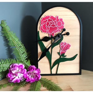 Peut inclure: Une horloge décorative en bois avec un cadre noir et un motif floral. Le cadran de l'horloge présente une fleur rose avec des feuilles vertes et des aiguilles noires. L'horloge est posée sur un fond en bois clair.