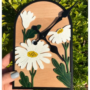 Peut inclure: Horloge murale en bois avec un motif de marguerites. Les marguerites blanches à cœur jaune sont entourées de feuilles vertes. Les aiguilles et le cadre sont noirs. Le fond est en bois clair, entouré de verdure.