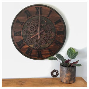 May include: A large, round wooden wall clock with Roman numerals and a gear mechanism in the center. The clock has a dark brown finish and is mounted on a white wall. A small potted plant and a decorative gear are on a wooden shelf below.