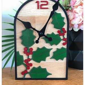 Peut inclure: Horloge décorative avec un cadre noir et un cadran en bois clair. Elle est ornée de feuilles de houx vertes et de baies rouges, avec le chiffre 12 en rouge. Les aiguilles sont noires.