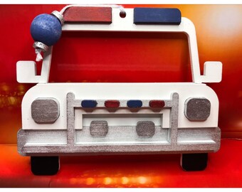 Può includere: Ornamento a forma di auto della polizia in legno bianco con dettagli rossi e blu. L'ornamento presenta una palla blu sulla parte superiore e dettagli color argento. Lo sfondo è un gradiente di rosso e arancione.