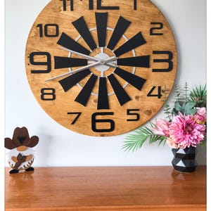 Könnte beinhalten: Eine große, runde Wanduhr aus Holz mit schwarzen Ziffern und Zeigern. Das Zifferblatt hat ein einzigartiges Design mit schwarzen geometrischen Formen, die vom Zentrum ausgehen. Eine kleine Zwergfigur und eine Vase mit rosa Blumen stehen auf einer Holzoberfläche.