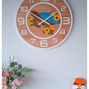 Könnte beinhalten: Eine runde Wanduhr aus Holz mit weißem Rahmen und großen weißen Zahlen. Das Zifferblatt zeigt ein Schildkröten-Design mit Sonnenblumen. Die Zeiger der Uhr sind schwarz. Die Uhr befindet sich an einer weißen Wand.