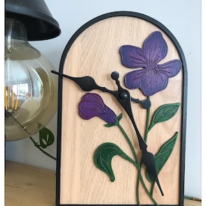 Peut inclure: Une horloge décorative en bois avec un motif floral. Le cadran est en bois clair, encadré de noir. Le motif floral comprend des fleurs violettes et des feuilles vertes. Les aiguilles de l'horloge sont noires.
