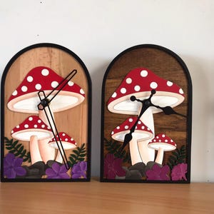 Peut inclure: Deux horloges décoratives sur le thème des champignons. Chaque horloge présente un chapeau de champignon rouge à pois blancs, un pied crème et un cadran noir. Les horloges sont posées sur un fond en bois et encadrées en noir.