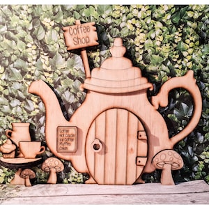 Könnte beinhalten: Holzschild für ein Café und eine türförmige Teekanne mit Pilz- und Kaffeetassendekor. Auf dem Schild steht "Coffee Shop". Die Tür hat ein kleines Schild mit Kaffee, heißer Schokolade, Eiskaffee, Muffins und Kuchen.