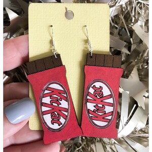 Könnte beinhalten: Ohrringe in Form von roten und braunen Kit Kat Schokoriegeln. Das Kit Kat-Logo ist weiß auf rotem Grund zu sehen. Die Ohrringe hängen an silbernen Haken und werden auf einer gelben Karte präsentiert.
