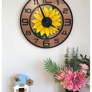 Peut inclure: Une horloge murale ronde en bois avec un cadre noir et de grands chiffres noirs. Le cadran de l'horloge présente un tournesol jaune avec des feuilles vertes. Un gnome et un vase de fleurs roses sont sur une surface en bois.