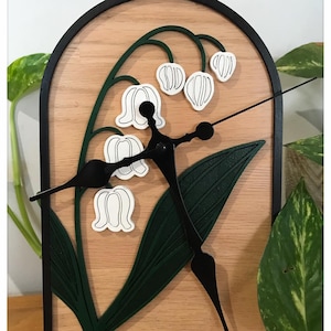 Peut inclure: Une horloge murale en bois avec un motif de muguet. Le cadran de l'horloge a un fond en bois clair avec un cadre noir en forme d'arche. Le muguet est blanc et vert, et les aiguilles de l'horloge sont noires.