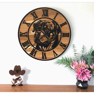 May include: A round wooden wall clock with a black Rottweiler dog design. The clock features black Roman numerals and hands. A small gnome figurine with a cowboy hat and a bouquet of pink flowers in a black vase are also visible.